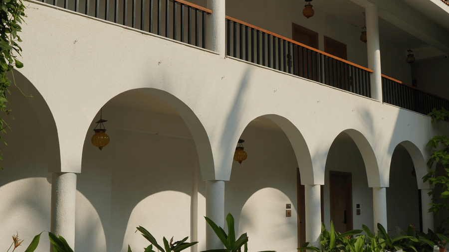 A white building exterior featuring large open arches, hanging lamps, and a balcony railing at Bougainvillea, Alibaug.