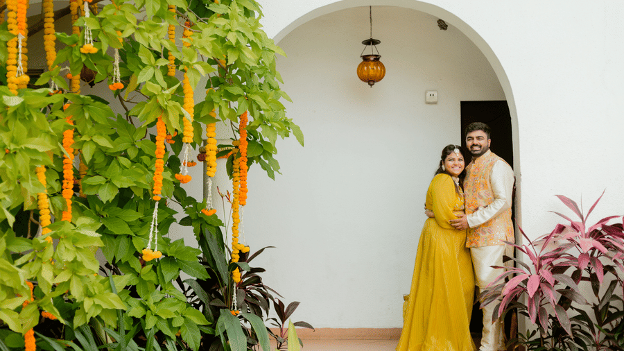 A couple posing together under an arched doorway decorated with floral garlands, leafy plants at Bougainvillea, Alibaug.