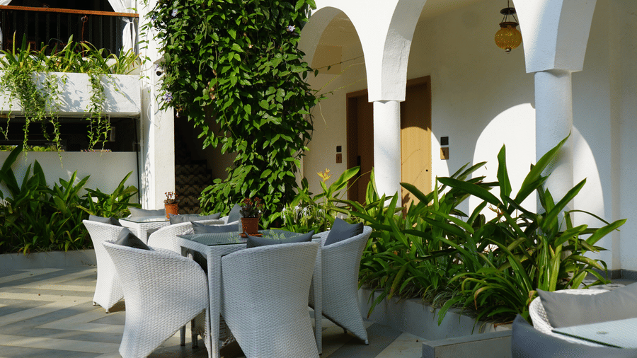 A white dining table surrounded by chairs, lush green plants, arched pillars at Bougainvillea, Alibaug.
