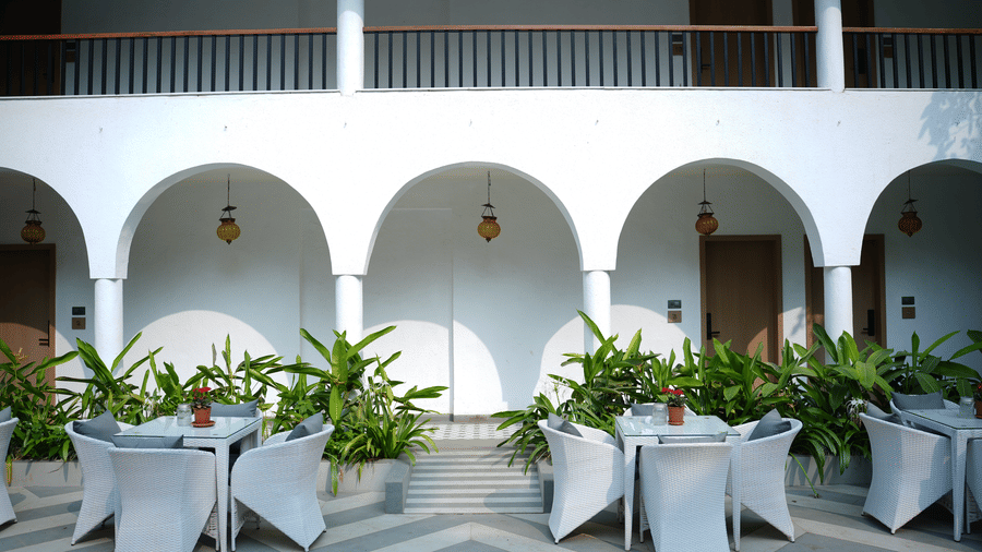 Dining sets are arranged on a chequered courtyard patio, backed by white arches, leafy potted plants at Bougainvillea, Alibaug.