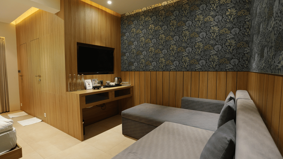 An L shaped sofa faces a wall mounted television unit with a wooden panel, patterned wallpaper, and warm lighting at Bougainvillea, Alibaug.