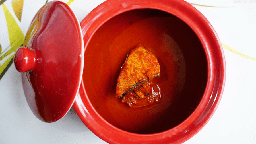 An aerial view of Goan fish curry served in a red claypot with the lit kept next to it.