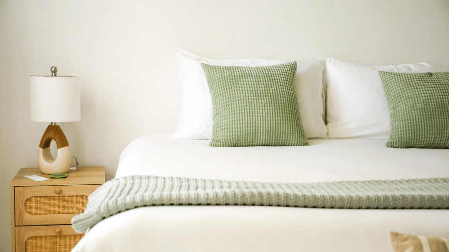 A bright, clean bedroom featuring a white bed with green throw pillows, a woven nightstand, and a modern lamp.
