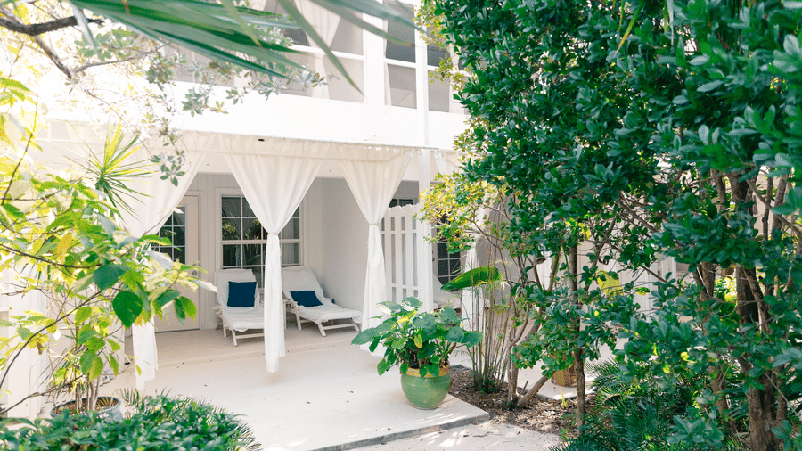 Two white lounge chairs with blue pillows sit on a white patio under a balcony, framed by white curtains and vibrant garden plants.