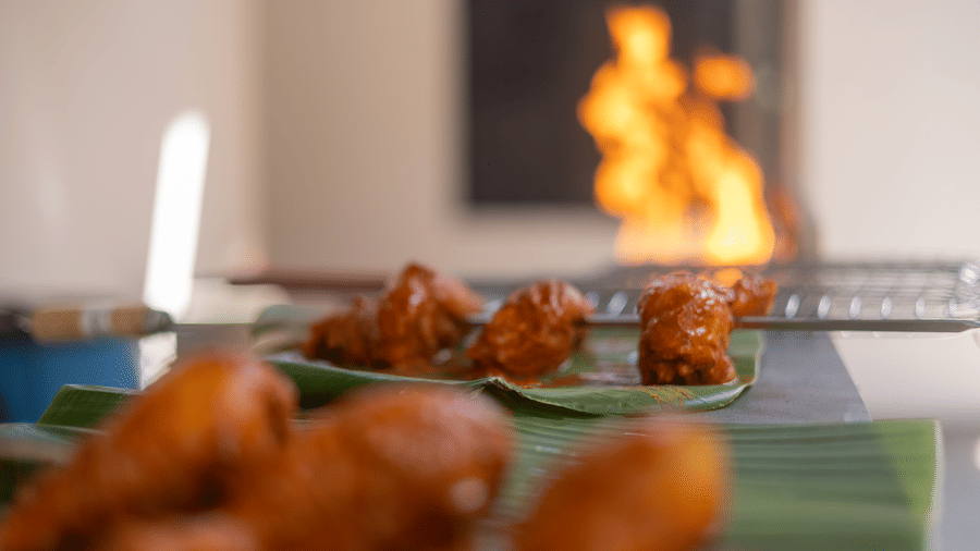 Close-up of cooked chicken pieces served on banana leaves with live kitchen flames roaring in the background.