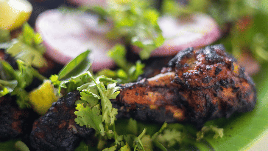 Close-up of charred grilled meat garnished with fresh herbs, sliced onions, and lemon wedges on a green leaf.