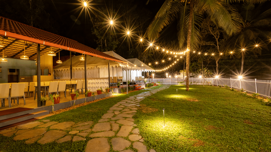 Paved stone pathway through a resort lawn at night, draped with glowing festoon lights overhead.