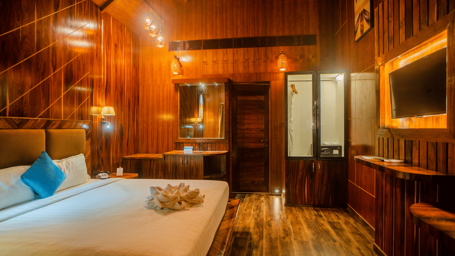 A wooden-themed hotel room featuring warm lighting, a comfortable bed with blue accent pillows, and wooden walls. The space has a rustic and inviting atmosphere.