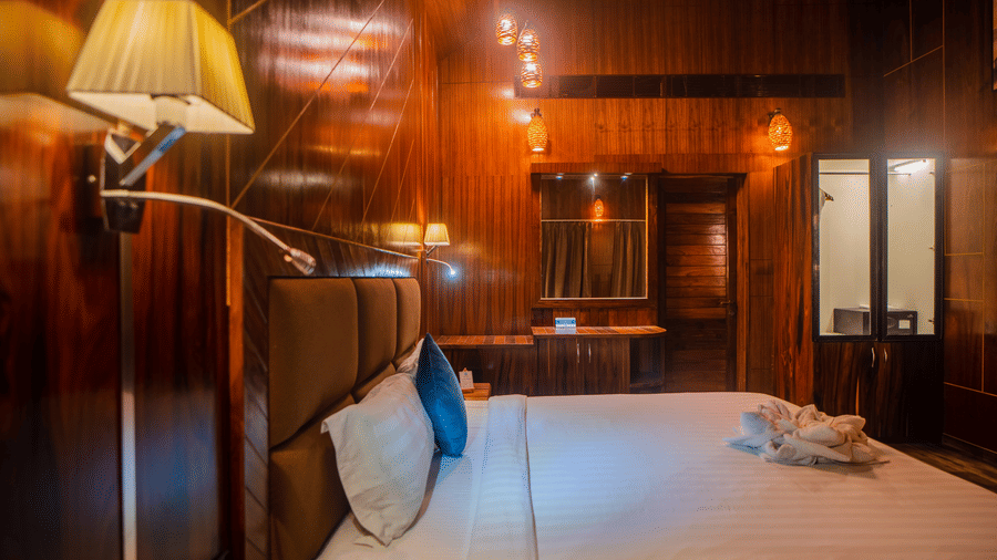  A well-lit hotel room with wooden walls and a bed adorned with blue accent pillows. The room also features wall lamps offering a relaxing vibe.