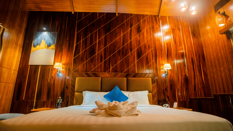A detailed shot of a wooden hotel room with a comfortable bed, warm lighting, and neatly arranged furnishings. The room has a peaceful charm.