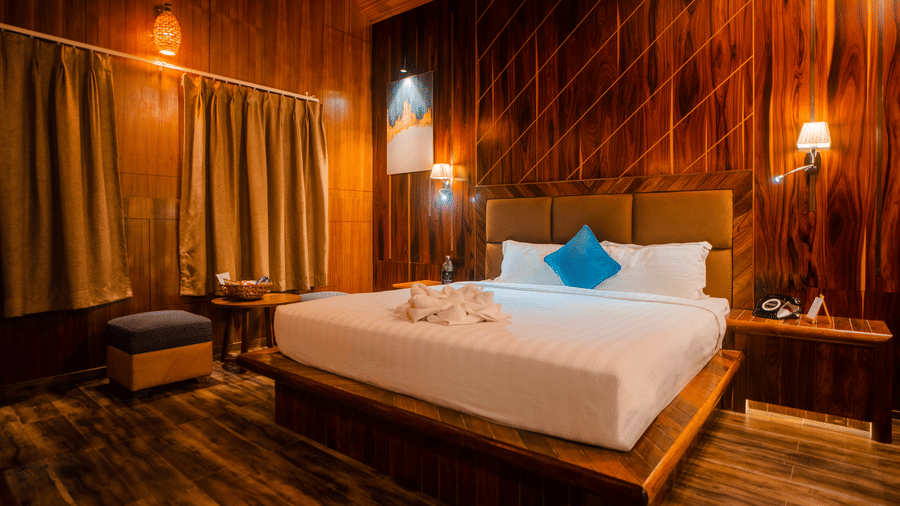  A cosy wooden hotel room featuring warm lighting, a large bed with blue accent pillows, and soft curtains. The wooden walls and  furniture create a comfortable, inviting atmosphere.