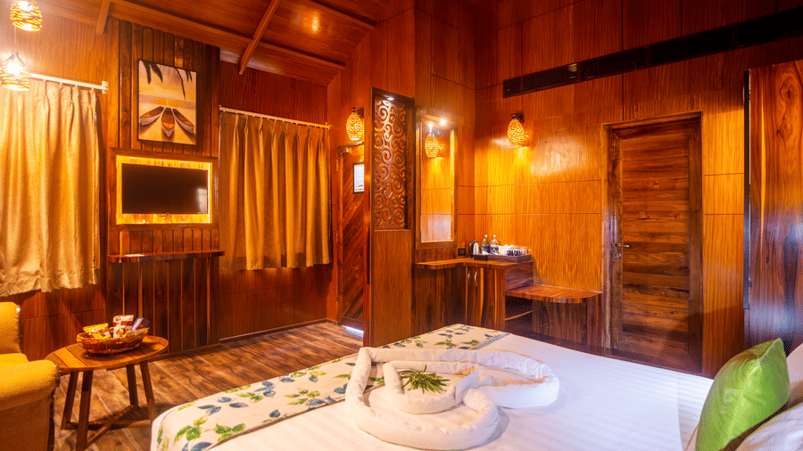 A warm, wooden hotel room featuring a large bed with green accent pillows, stylish lighting, and a small table with decorative items. The room's design is complemented by the ambient lighting.