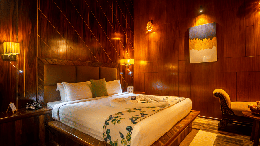 A wooden-themed hotel room featuring a comfortable bed with white linens and green accent pillows. The room has warm lighting, a cosy atmosphere, and rustic wooden walls.