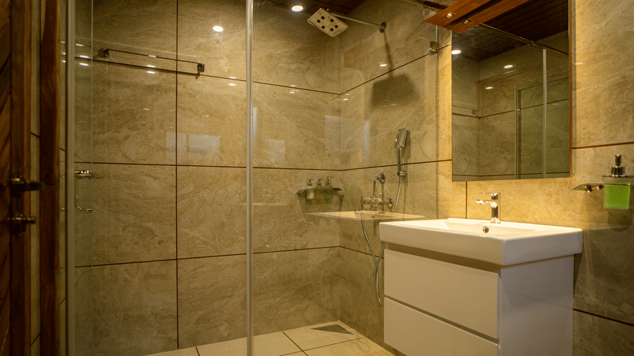 A spacious bathroom with a walk-in shower, large tiles, and a sleek white sink. The soft lighting and natural light from the window make the space feel open and airy.