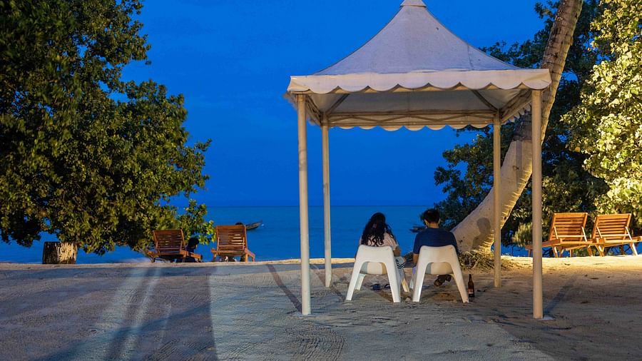 A tranquil beach scene with a couple sitting on lounge chairs near the shore, overlooking the calm sea. The sky is softly lit by the evening or morning sun - Coral Reef Resort & Spa, Havelock