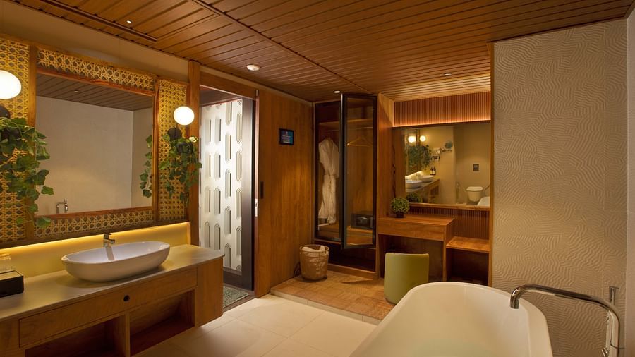 Bathroom of Coral Teal Suite at Coral Reef Resort and Spa in Havelock featuring a sink with a mirror over it and lamps on the both sides opposite to which is a bathroom and in another corner is a dressing mirror with a pouffe