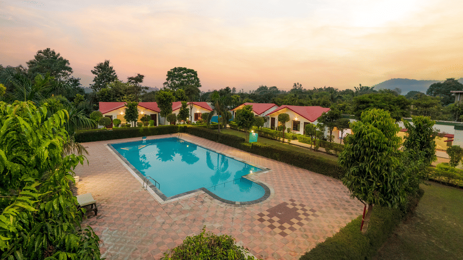 An aerial view of the swimming pool at Corbett Holiday Forest Resort, Ramnagar, during the evening hours with a bright orange sky in the background.