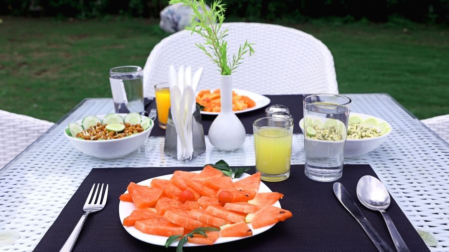 A neatly arranged outdoor dining table is set with fresh fruit, beverages, and cutlery at Corbett Nirvana Resort.