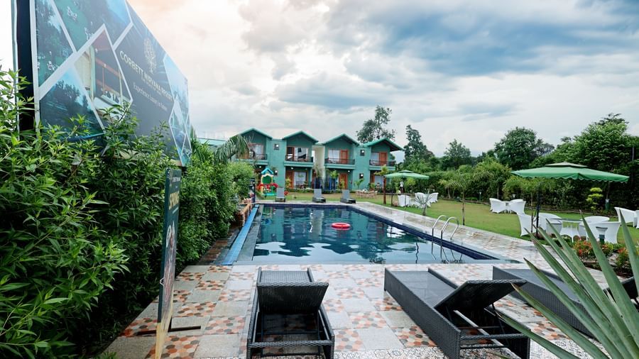 Guests can relax on sun loungers by the pool with a view of the modern resort buildings at Corbett Nirvana Resort.