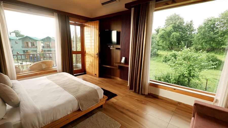 Spacious room with large windows and balcony offering lush greenery views at Corbett Nirvana Resort.