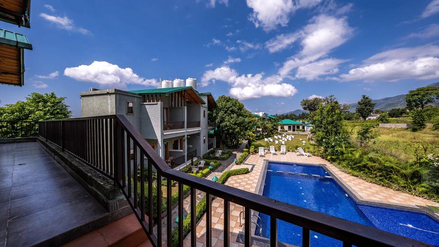 A closeup shot of verandah at Corbett Nirvana Resort, and a view of two storey building, a swimming pool, a lawn with seating arrangements, and some cottage style buildings at a distance.