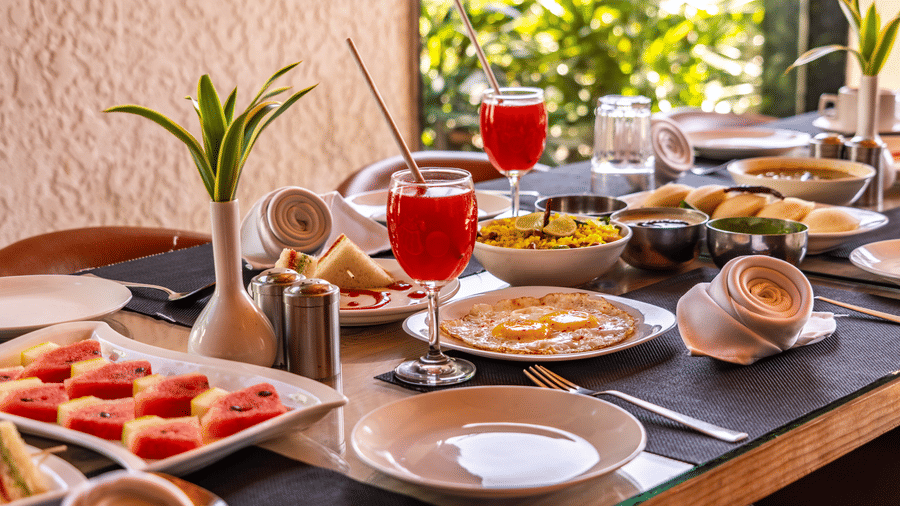 Close-up view of a dining table at Corbett Nirvana Resort set with an assortment of dishes, including plates of food, fruit, and drinks.