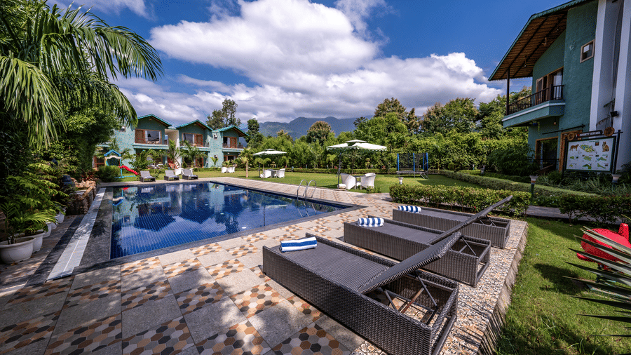 Day-time view of the swimming pool area at Corbett Nirvana Resort, showing modern lounge chairs on a deck, with foliage and the resort buildings in the background.