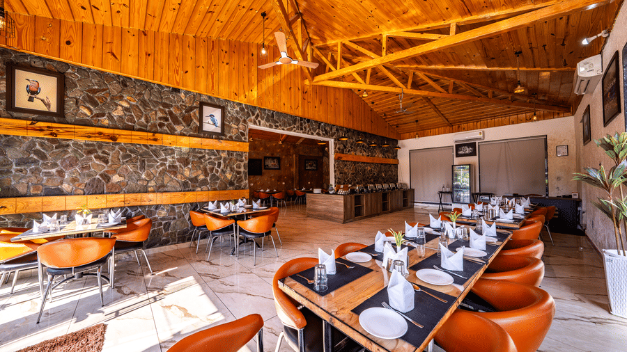 Interior view of the restaurant at Corbett Nirvana Resort with rustic stone accents, wooden walls, and long dining tables with orange chairs.