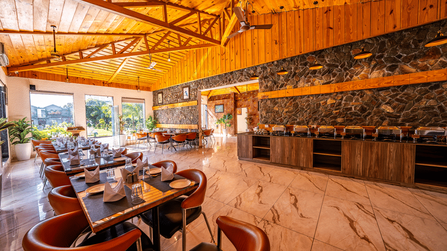 Wide interior shot of a spacious restaurant at Corbett Nirvana Resort with a high wooden ceiling, orange chairs, and a buffet serving counter.
