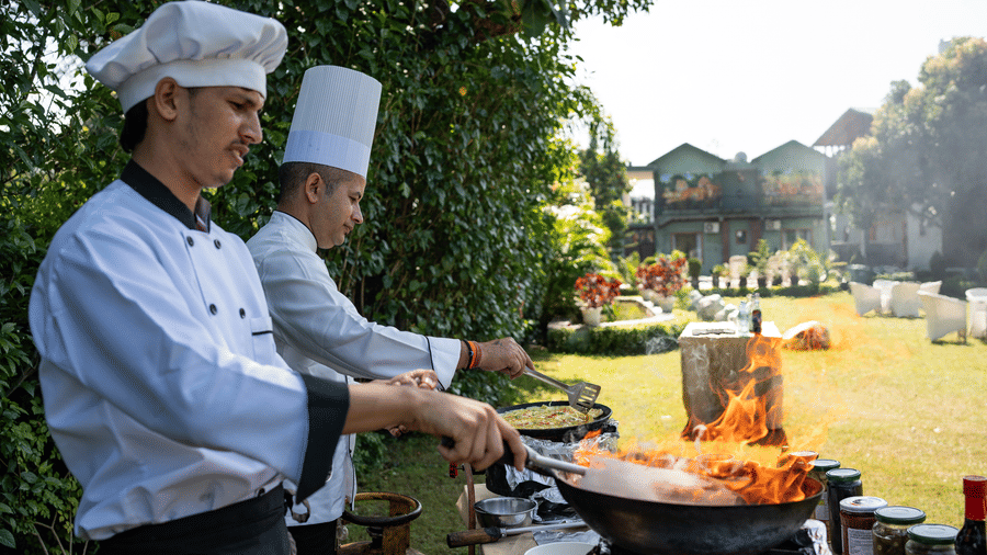 Two chefs in white hats preparing food outdoors over a fire in a large wok at Corbett Nirvana Resort, suggesting an outdoor catering or live cooking station.
