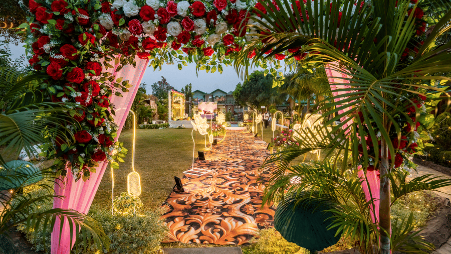 A well-lit pathway and decorated archway leading through the garden area of Corbett Nirvana Resort at dusk, with lush greenery and floral arrangements.