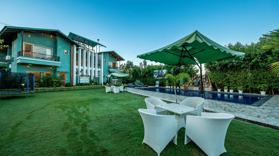 Wide-angle day-time view of the pool area and lawn at Corbett Nirvana Resort, showing the teal-coloured building and white outdoor seating.