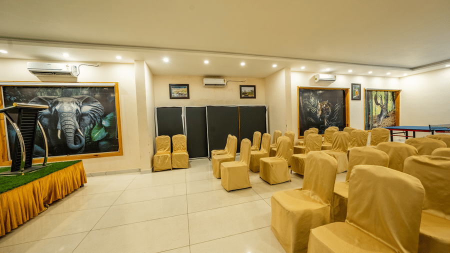 Banquet Hall at Corbett Nirvana Resort featuring several sets of tables and chairs in a classroom setting, a stage with podium and white board, and a few ACs installed. 