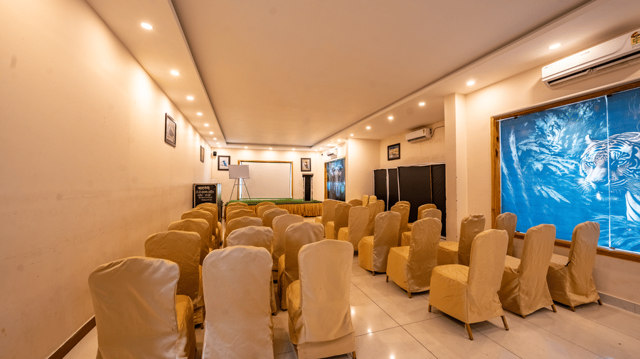 Banquet Hall at Corbett Nirvana Resort featuring several sets of tables and chairs in a classroom setting, a stage with podium and white board, and a few ACs installed. 