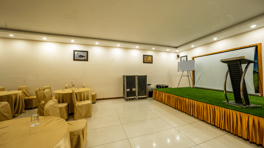 Banquet Hall at Corbett Nirvana Resort featuring several sets of tables and chairs in a classroom setting and a stage with podium and white board. 