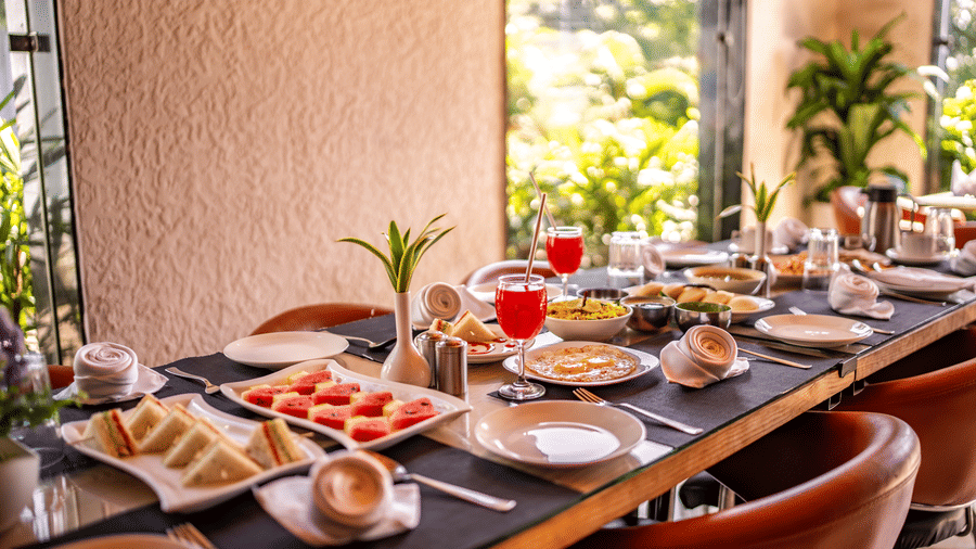 A dining table at Corbett Nirvana Resort is set with plates of sandwiches, fruit, and drinks next to a window with plants outside.
