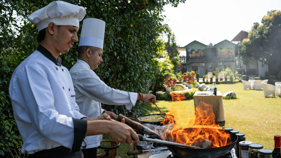 Corbett Nirvana Resort featuring two chefs in uniforms cook food in a pan over a flame at an outdoor station with buildings and trees behind them.