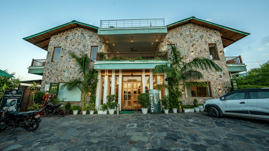 Facade of Corbett Nirvana Resort, Uttarakhand featuring the two storey building with balconies and terrace, a driveway before the entrance where vehicles are parked and there are few plants and flower pots nearby.