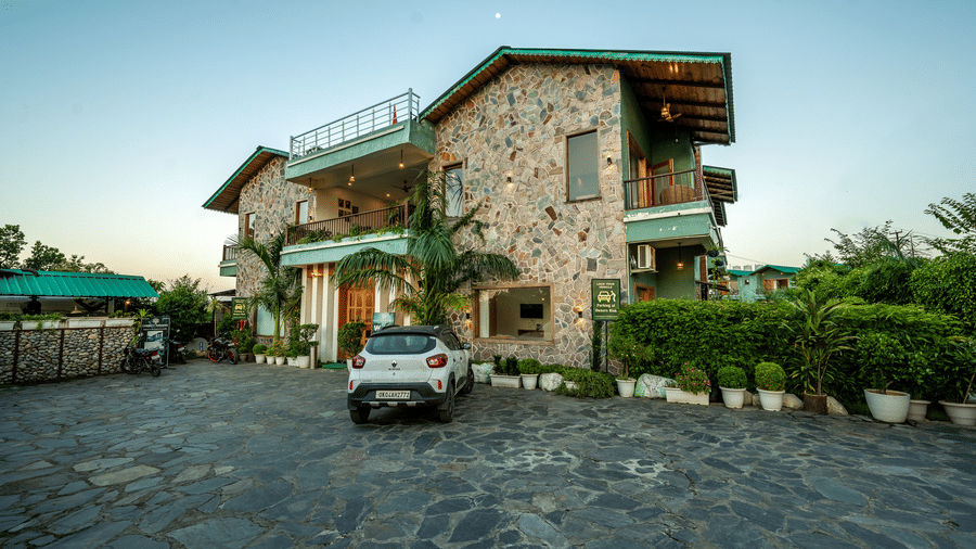 Facade of Corbett Nirvana Resort, Uttarakhand featuring two storey cottage style building and a huge concrete lawn where vehicles are parked.