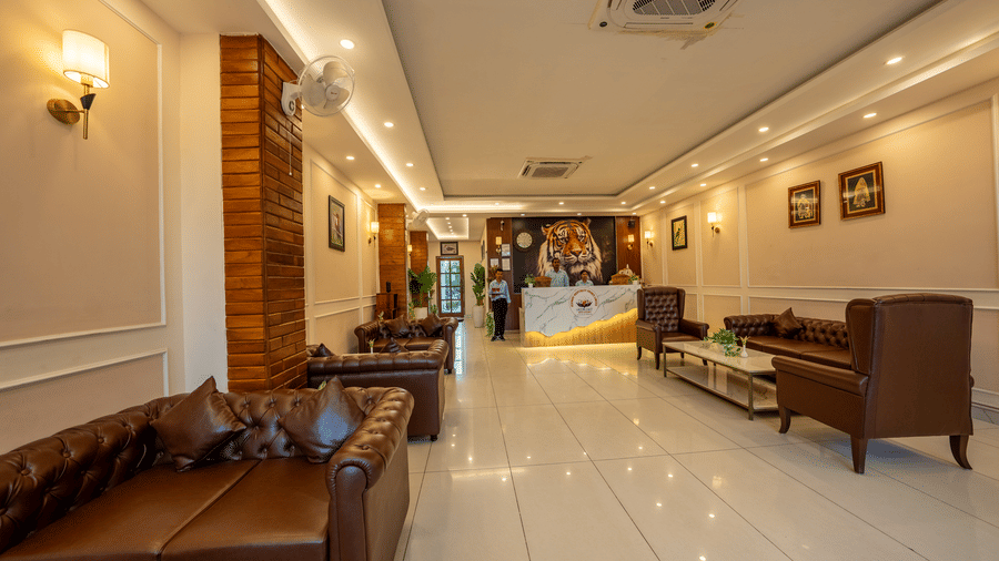 Sofas and chairs are arranged in a lobby with a reception desk and wall lights at the Corbett Nirvana Resort.