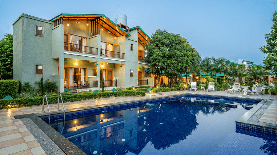 Outdoor swimming pool with tiled deck, lounge chairs, and a two-storey building with balconies and lights turned on, surrounded by trees at Corbett Nirvana Resort.