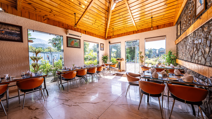An indoor dining room at Corbett Nirvana Resort featuring tables and chairs set for service under a vaulted ceiling with windows looking out to greenery.