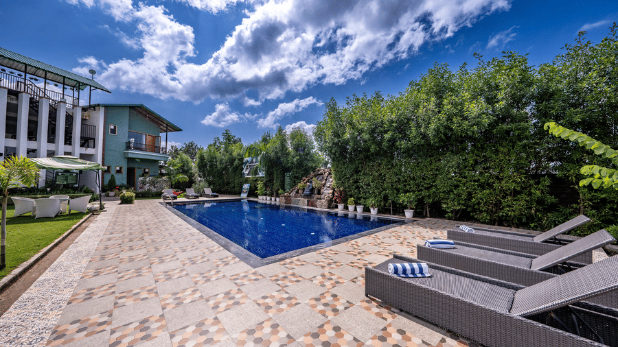 A swimming pool at Corbett Nirvana Resort surrounded by trees, lawn, and  facade buildings. 