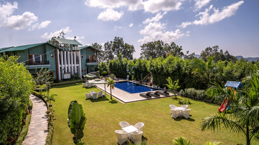 A swimming pool at Corbett Nirvana Resort surrounded by trees, facade building, and a huge lawn with seating arrangements.
