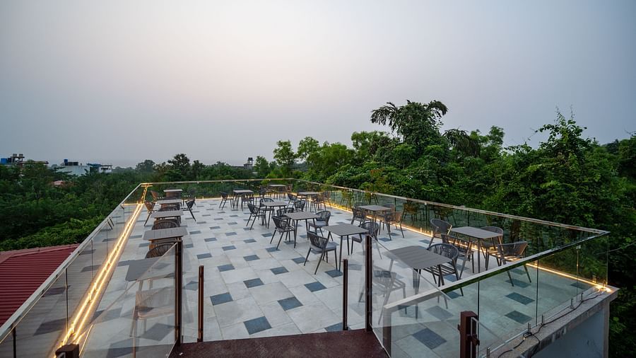 A rooftop cafe/bar featuring chairs and tables arranged within a glass railing, with lush greenery forming a scenic backdrop - Crescent Resort, Goa.