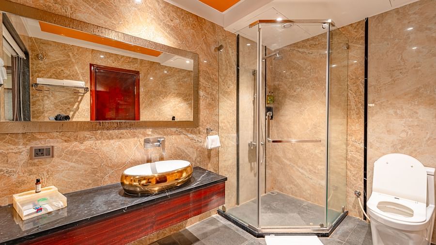 Deluxe Room bathroom features a rectangular mirror above the wash basin, a shower head to the right, a wall-mounted hand wash to the left, and a glass panel separating the shower area at Crescent Spa & Resort, Indore