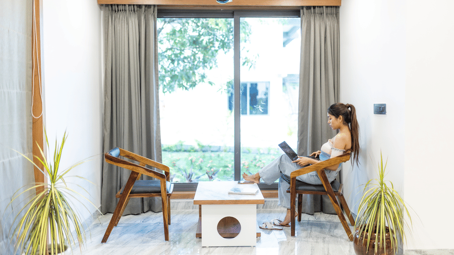 A woman sitting in the seating area for 2, located by the large glass windows with curtains.