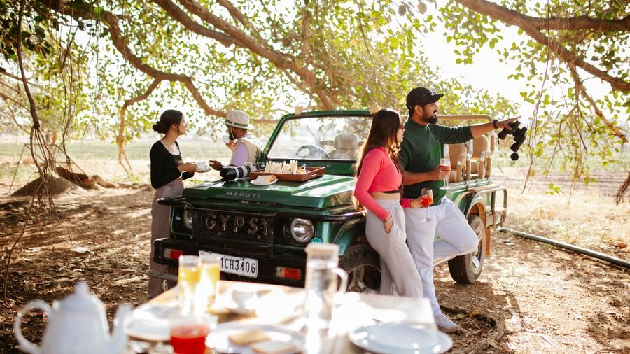 Four people standing beside a jeep which is standing before a breakfast table, surrounded by trees -Daksh Eden Greenz, Sasan Gir