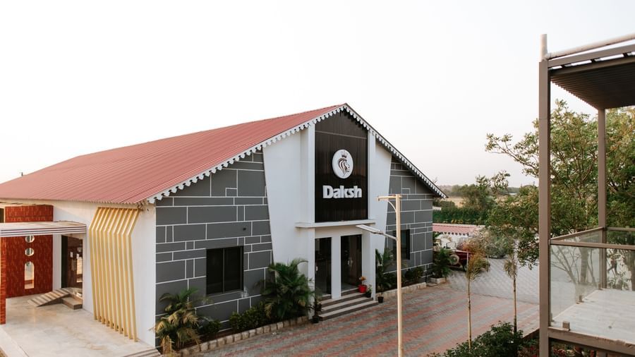 A side angle shot of a the exterior of Daksh Eden Greenz, Sasan Gir on a bright day, surrounded by trees and spacious walkways.