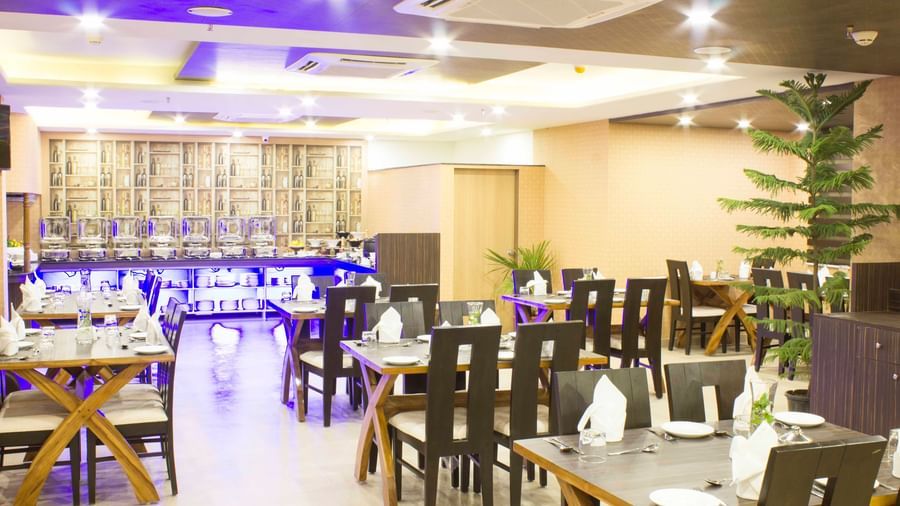A hotel restaurant with set tables, dark chairs, and blue ambient lighting | Daksh The Madhuvan Suites